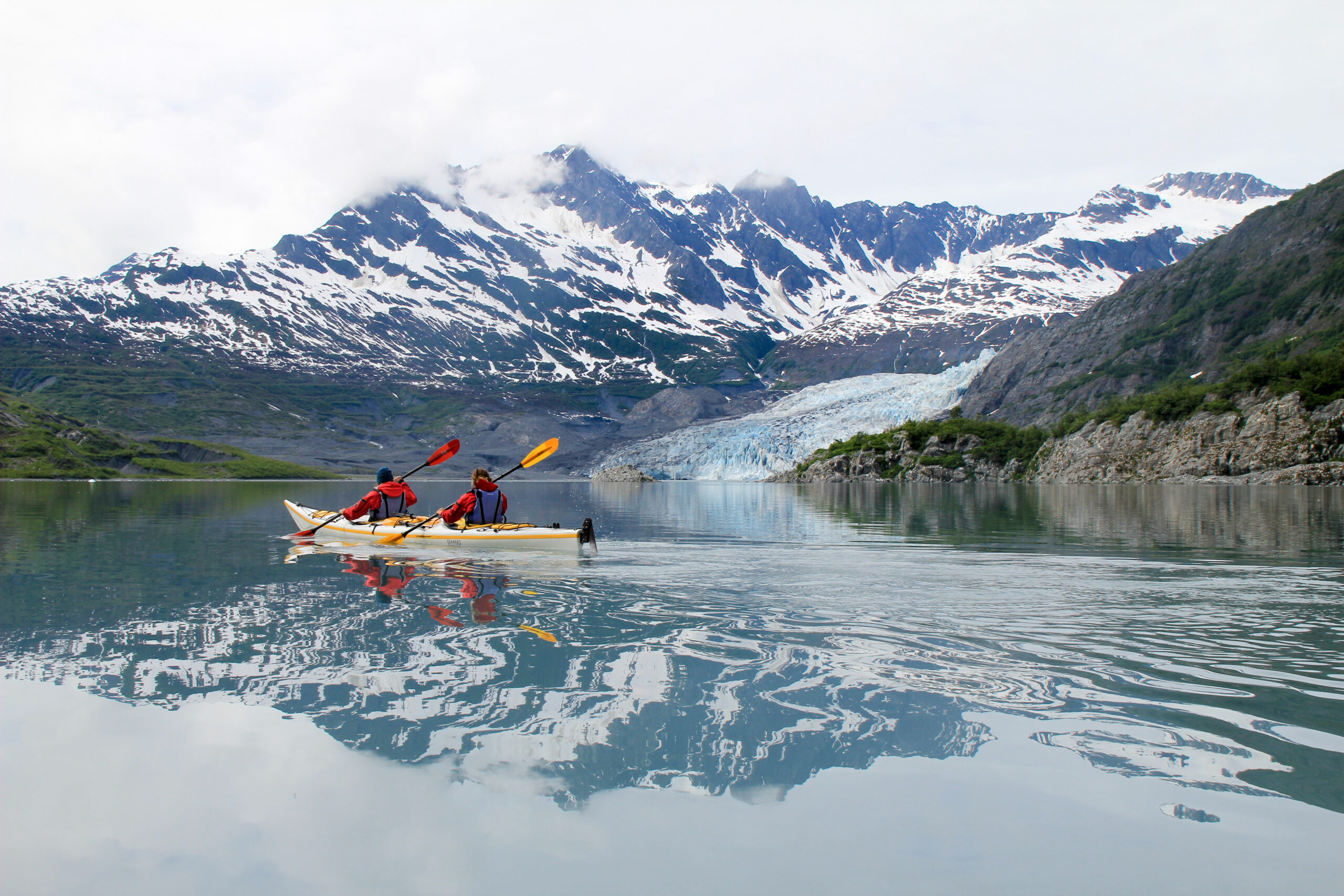 two people in a tandem sea kayak paddling across a lake with snowcapped mountains in the background