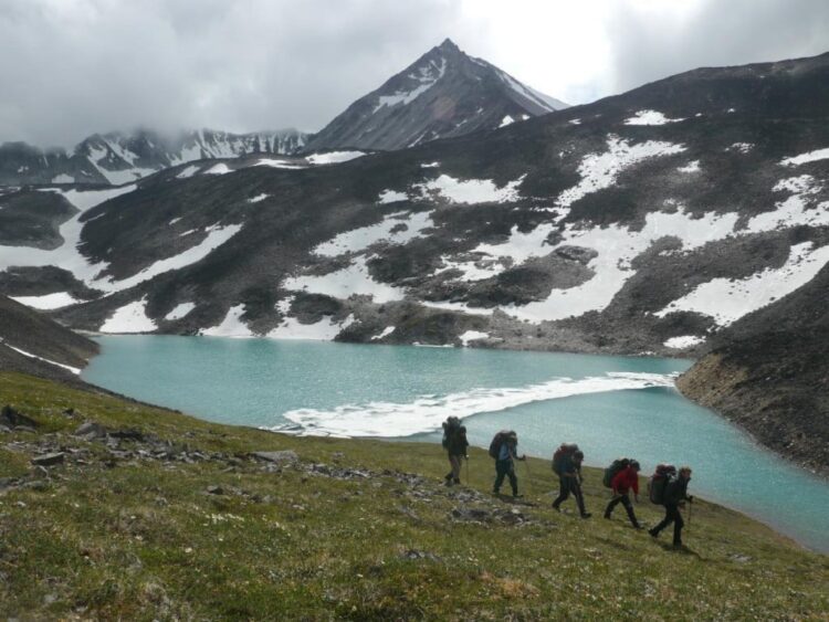Group of teens backpacking through alaska at the base of a mountain with a lake and snow behind them