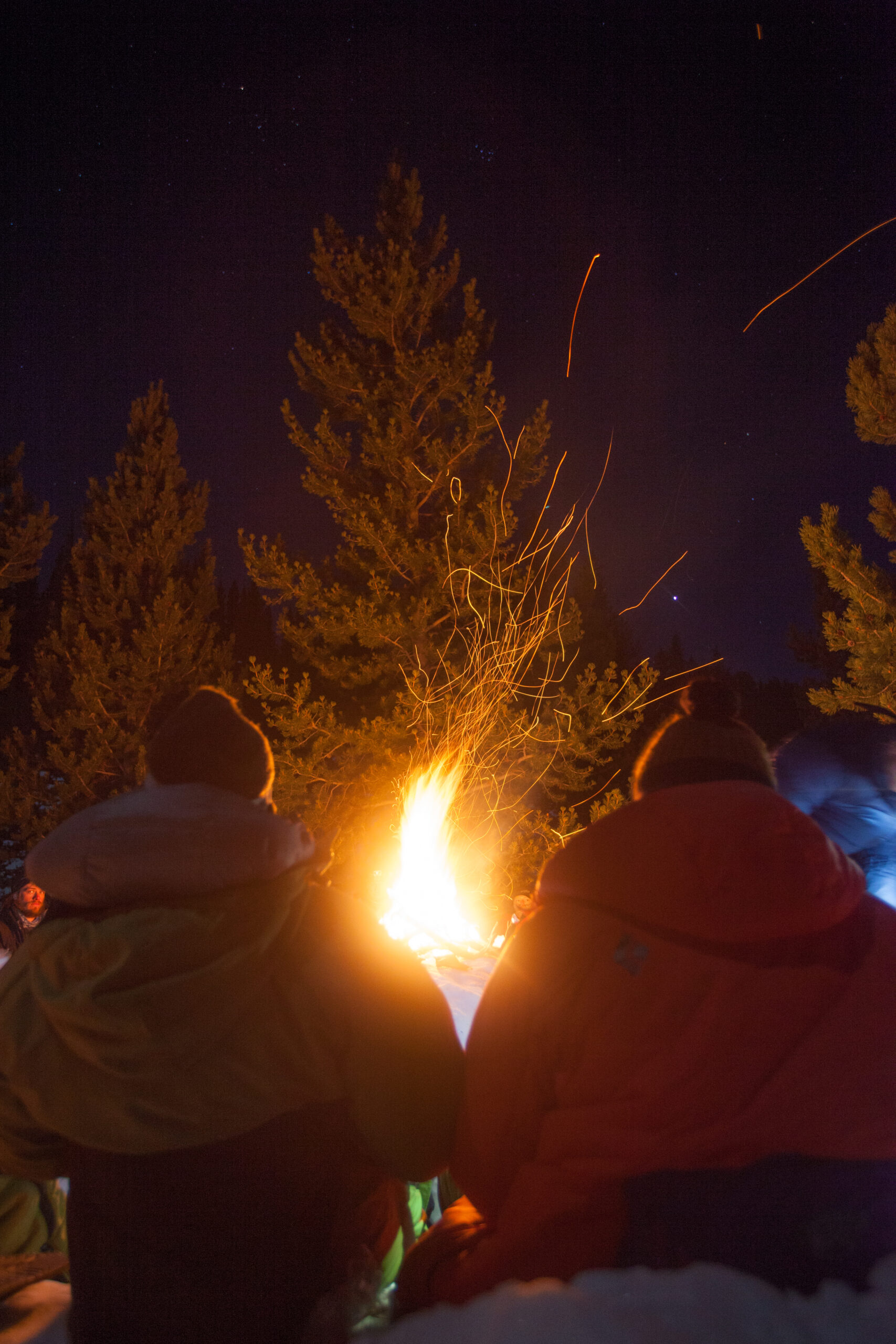 Two people sit by a fire. The sky is dark and there is snow on the ground