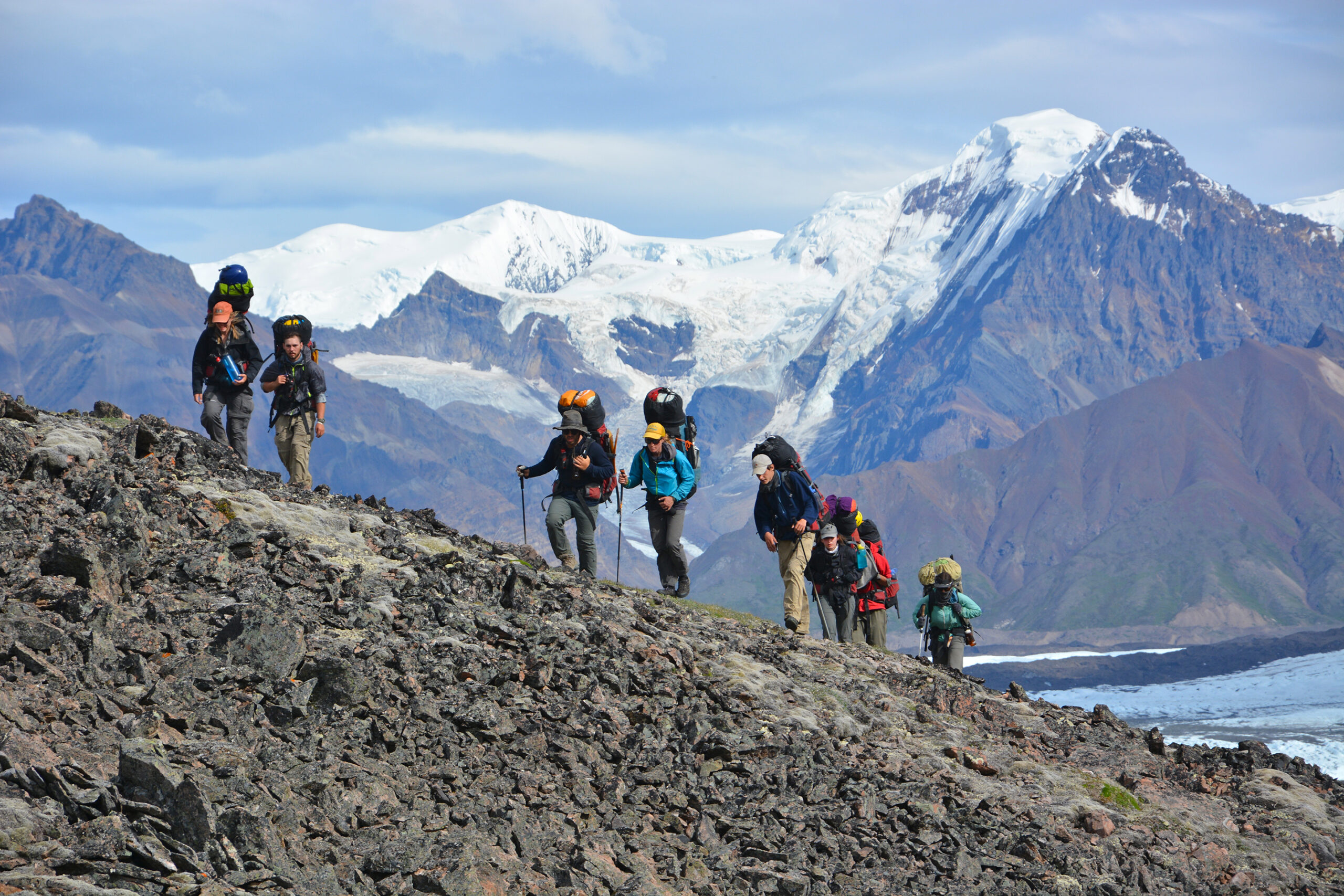 Group of adults backpacking up a rocky incline with snow capped mountains in the background