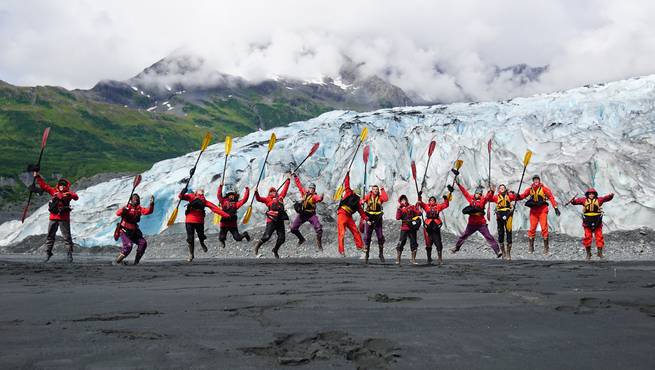 A course jumps together with their sea kayaking paddles on a beach in Alaska.