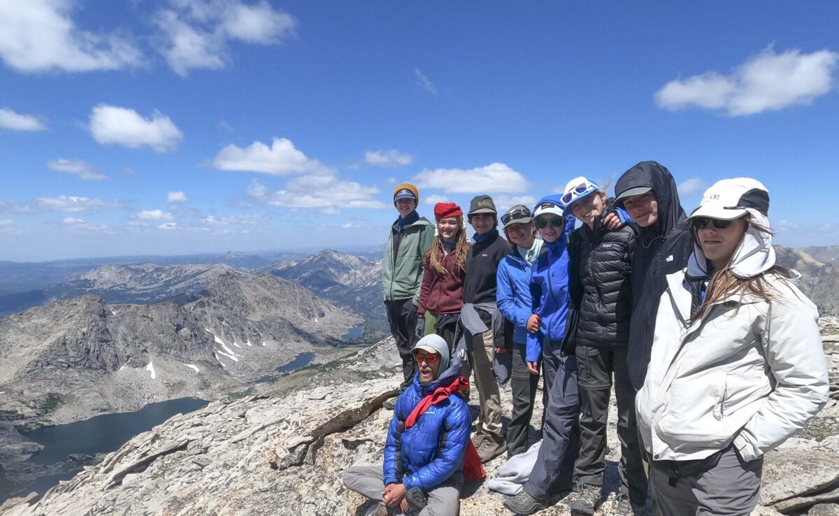 Group of teens standing on top of a large rock at elevation with mountains off in the background