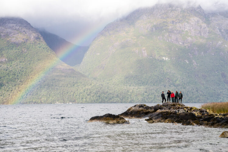 a group of people stand on a rock in the ocean. There is a rainbow behind them and mountains in the background.