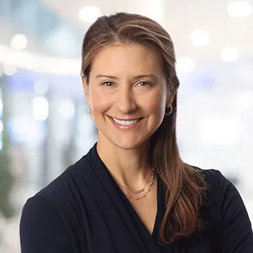 A woman with medium-length brown hair poses against a blurry background. She is smiling at the camera.