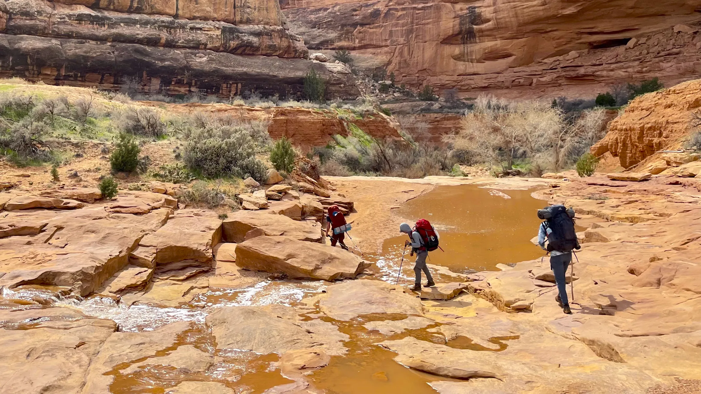 Three people wearing backpacking backpacks walk on red rocks. There are tall red canyon walls in the background.