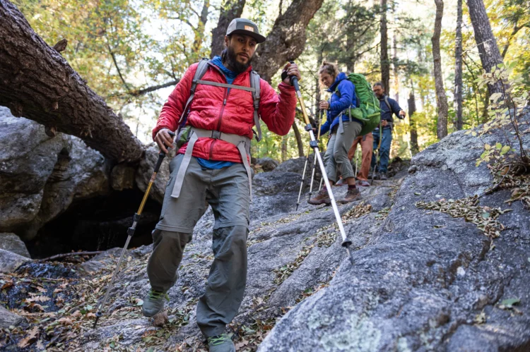 A man in a red jacket wearing a backpack and using trekking poles hikes down a granite rock. There are two women hikers on the trail behind him.