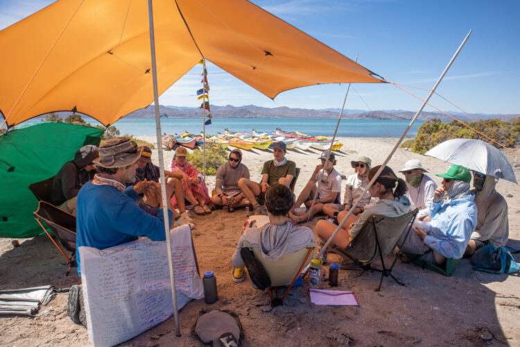 A group of people sit on a sandy beach under a yellow tarp.
