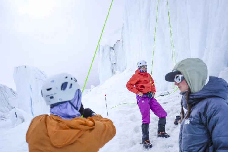 A woman in warm clothing and a climbing hardness stands at the base of an ice wall. She is smiling at a person in the foreground.