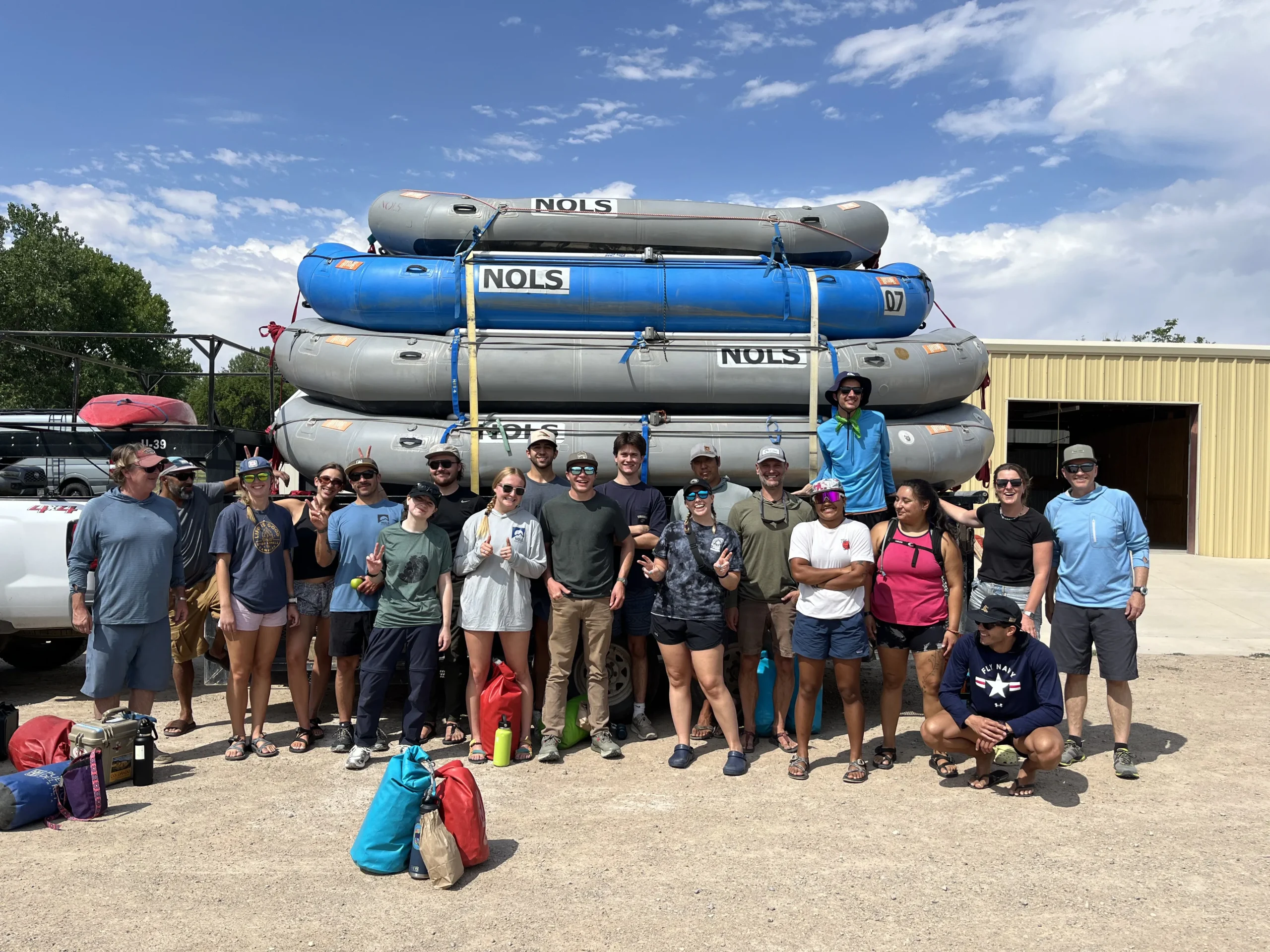 A large group of people pose in front of a trailer piled with river rafts. It's sunny and looks warm based on their clothing.