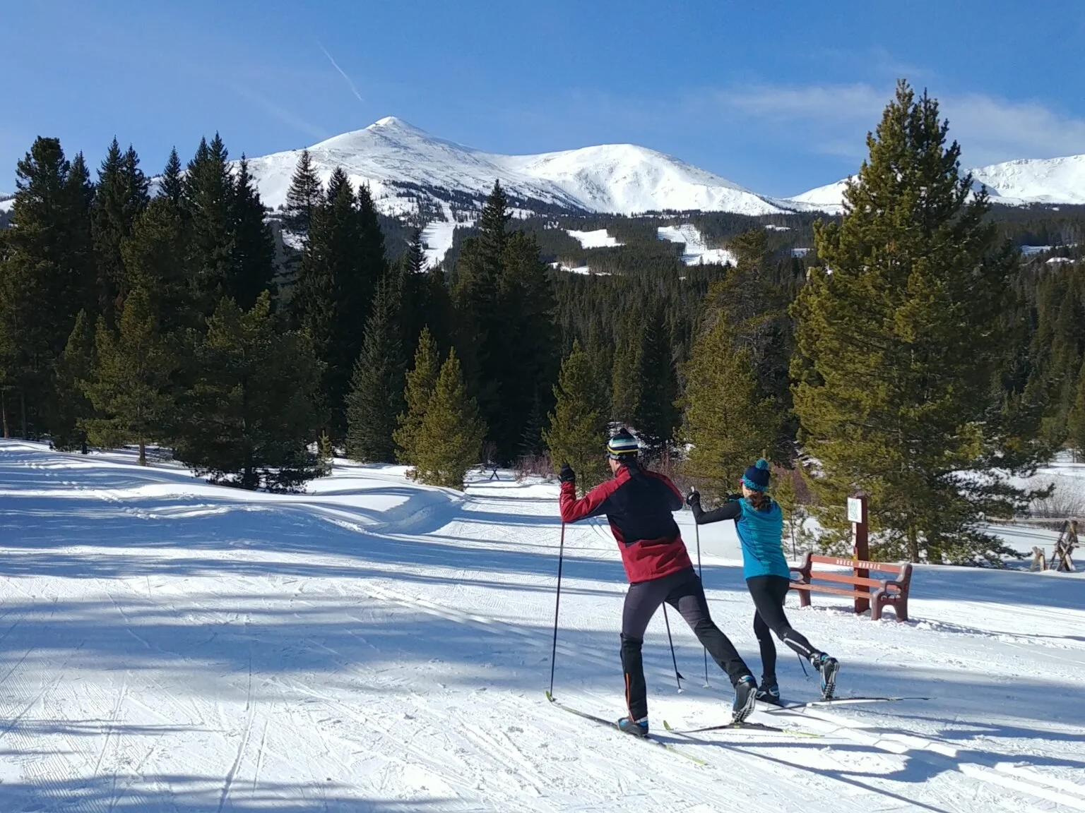 Two cross-country skiers on a trail with snow covered-mountains in the background.
