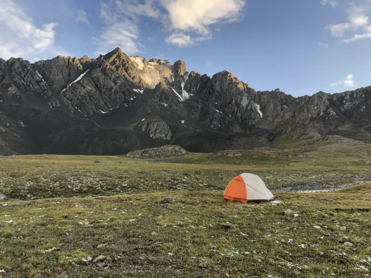 A backpacking tent sits in a rocky grassy field. There are sharp mountains in the background.