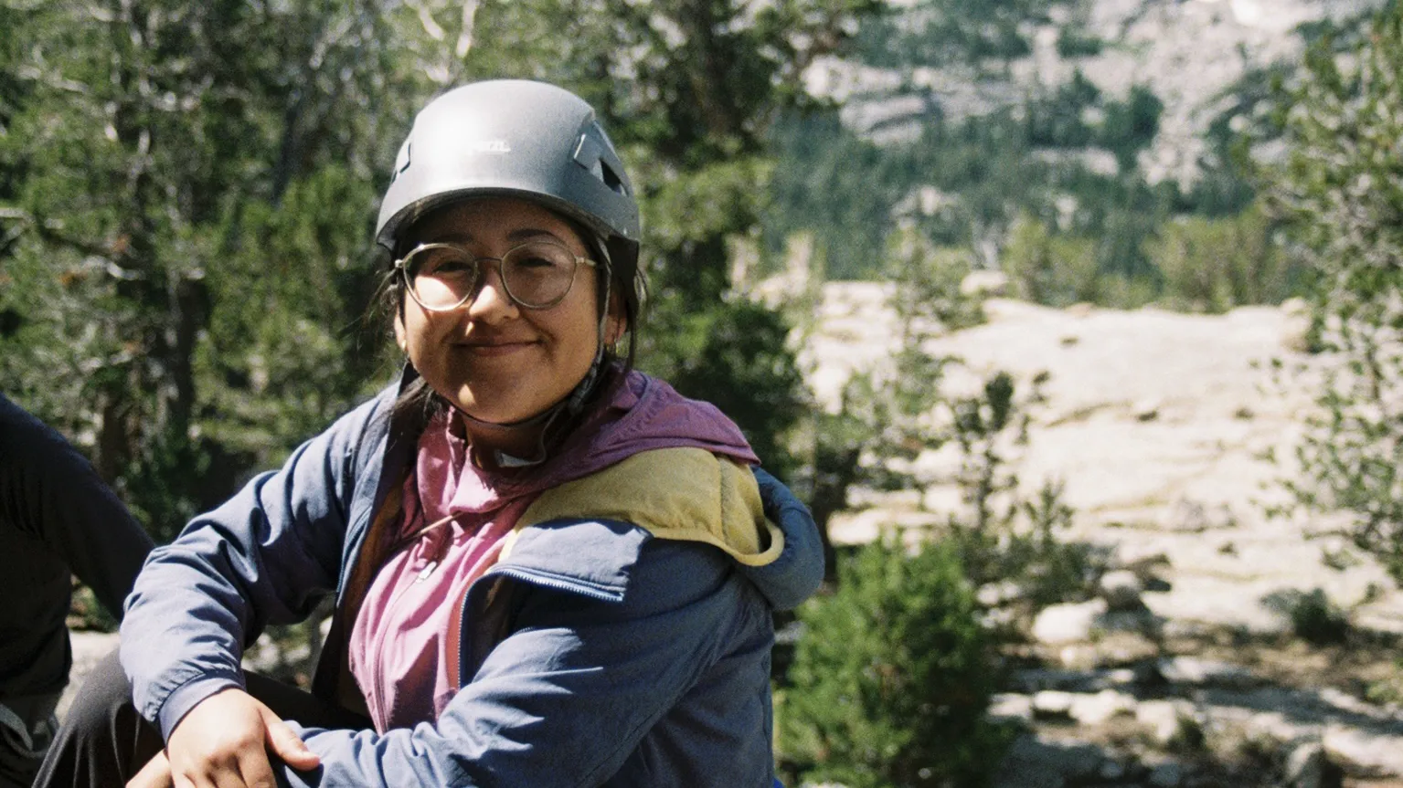A woman wearing a climbing helmet and glasses smiles at the camera. She has dark brown hair that's tucked behind her jacket hood. In the background are pine trees, granite rocks, and mountains.