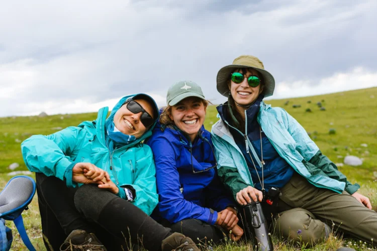 Three young women in rain jackets and sun hats sit together on an open grassland, laughing and leaning into each other, with rolling green hills and stormy skies in the background.