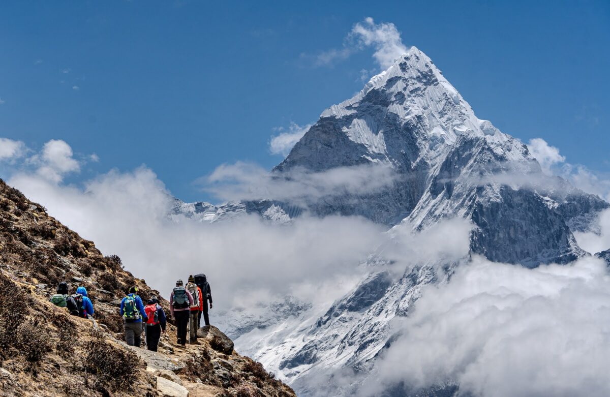 Six members of the team, photographed from behind, on a path with filmy clouds and Everest in the background.