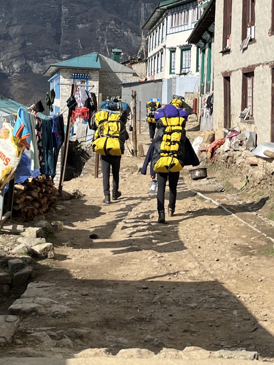 Members of the team, shown from behind, walk through a small town on a dirt road.