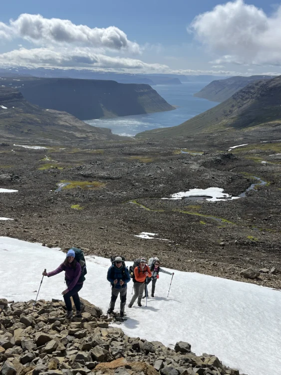a group of people hike up a small snow field on a rocky field on a ridge above the ocean.