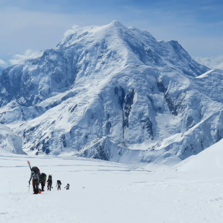 A group of mountaineers with large backpacks travel in a line up a snow field. A very tall mountain looms in the background.