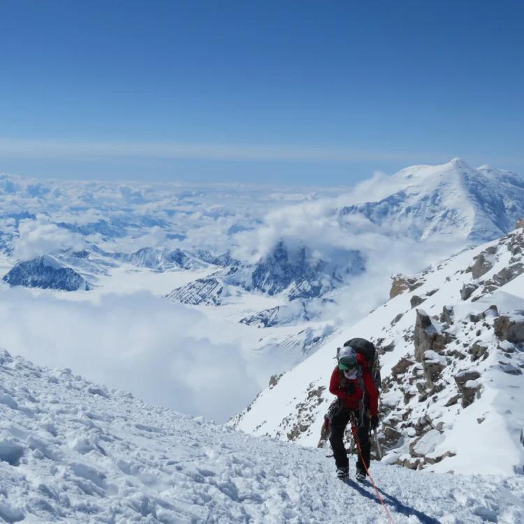 A mountaineer stands on a snowy ridge. There are endless mountains in the background.