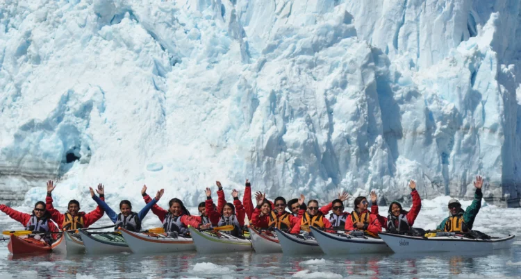 A large group of people in sea kayaks pose with arms up in front of a large glacier.