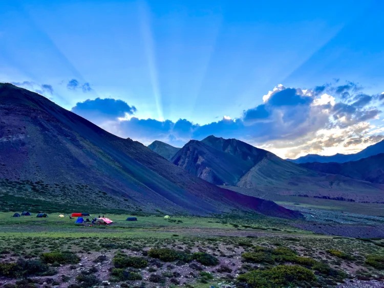 The sun sets behind blue mountains with tents set up on the valley.