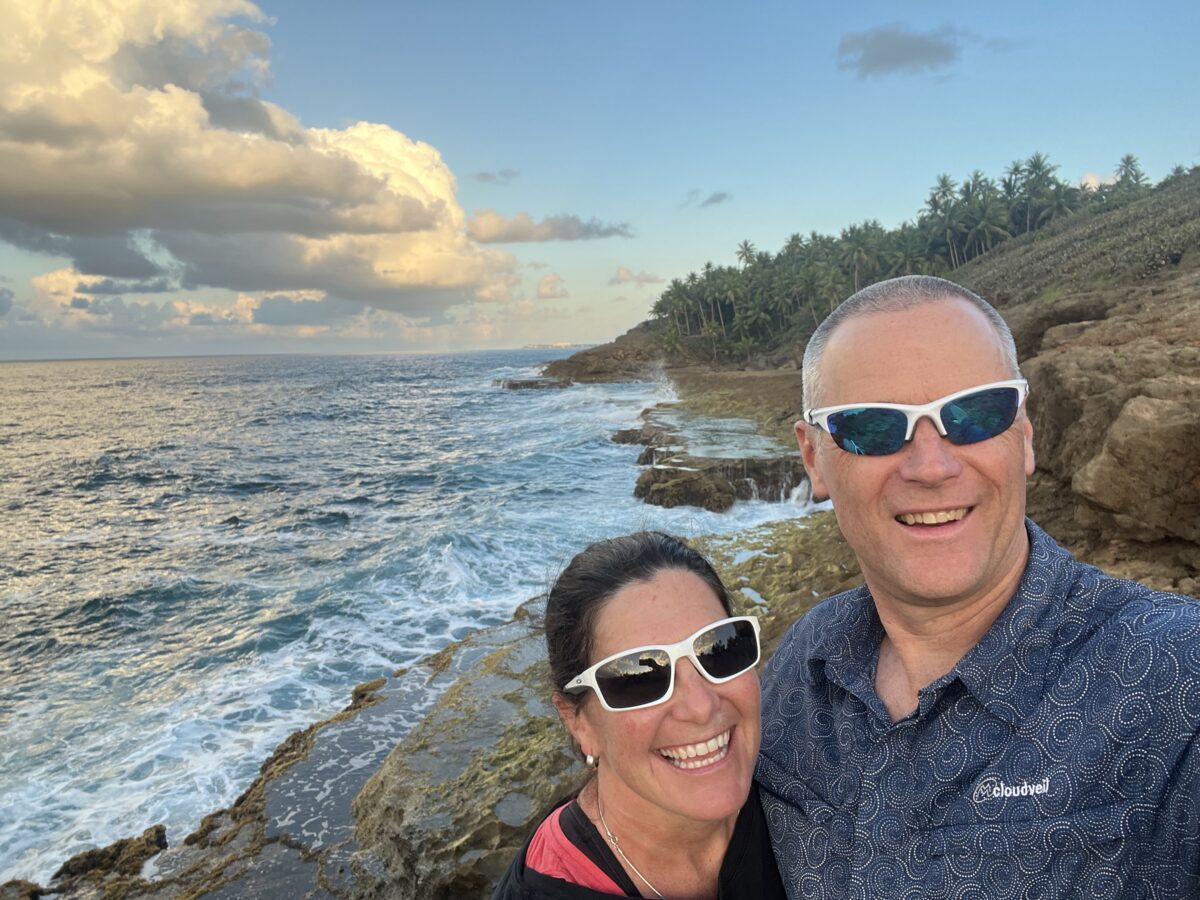 Clair Parrish Smith and Steve Smith, both wearing sunglasses, stand close together by the ocean on a rocky cliff with blue skies and clouds in the background.