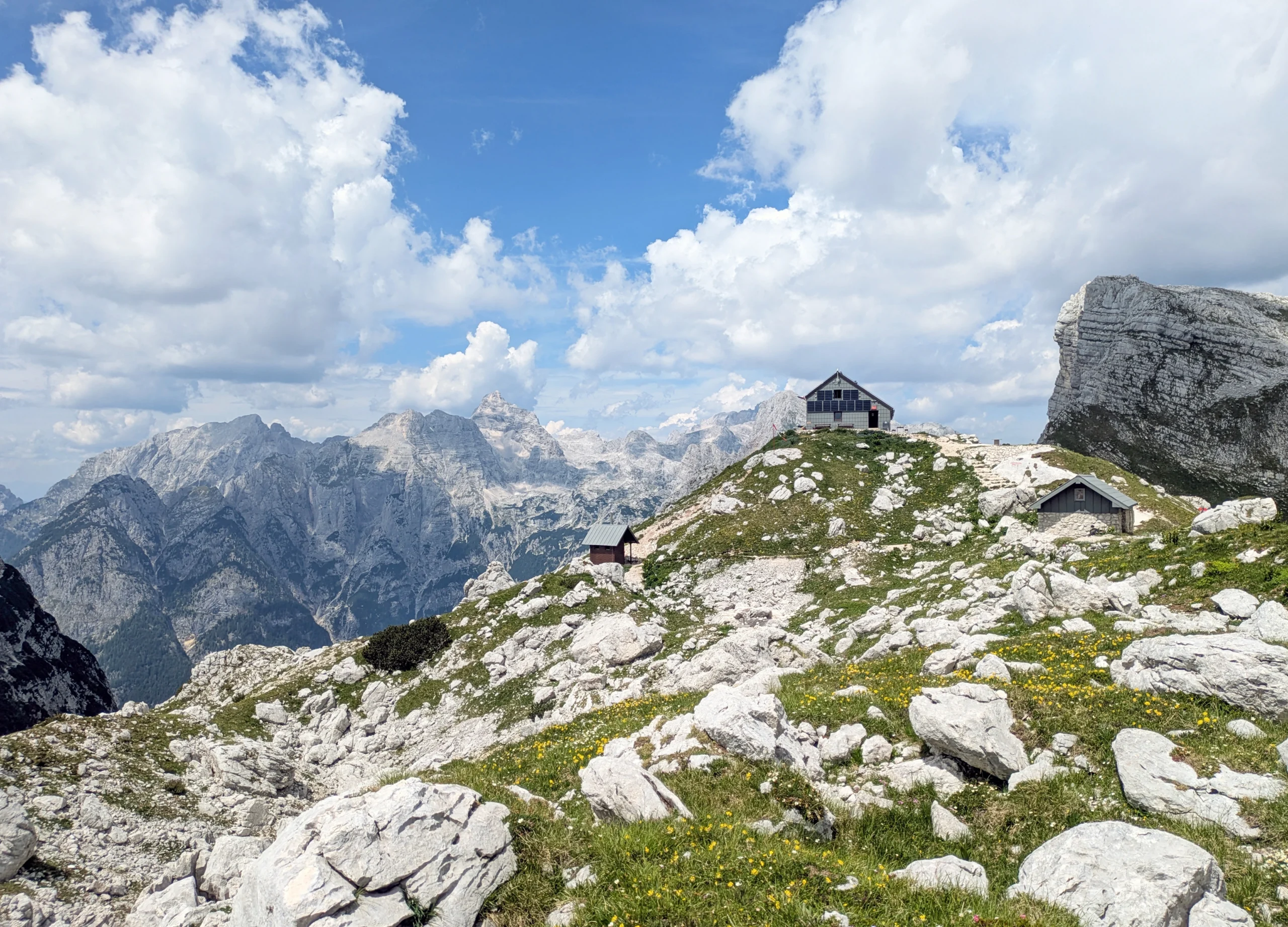 Mountain hut sits atop rocks and grass with dramatic mountains in the background