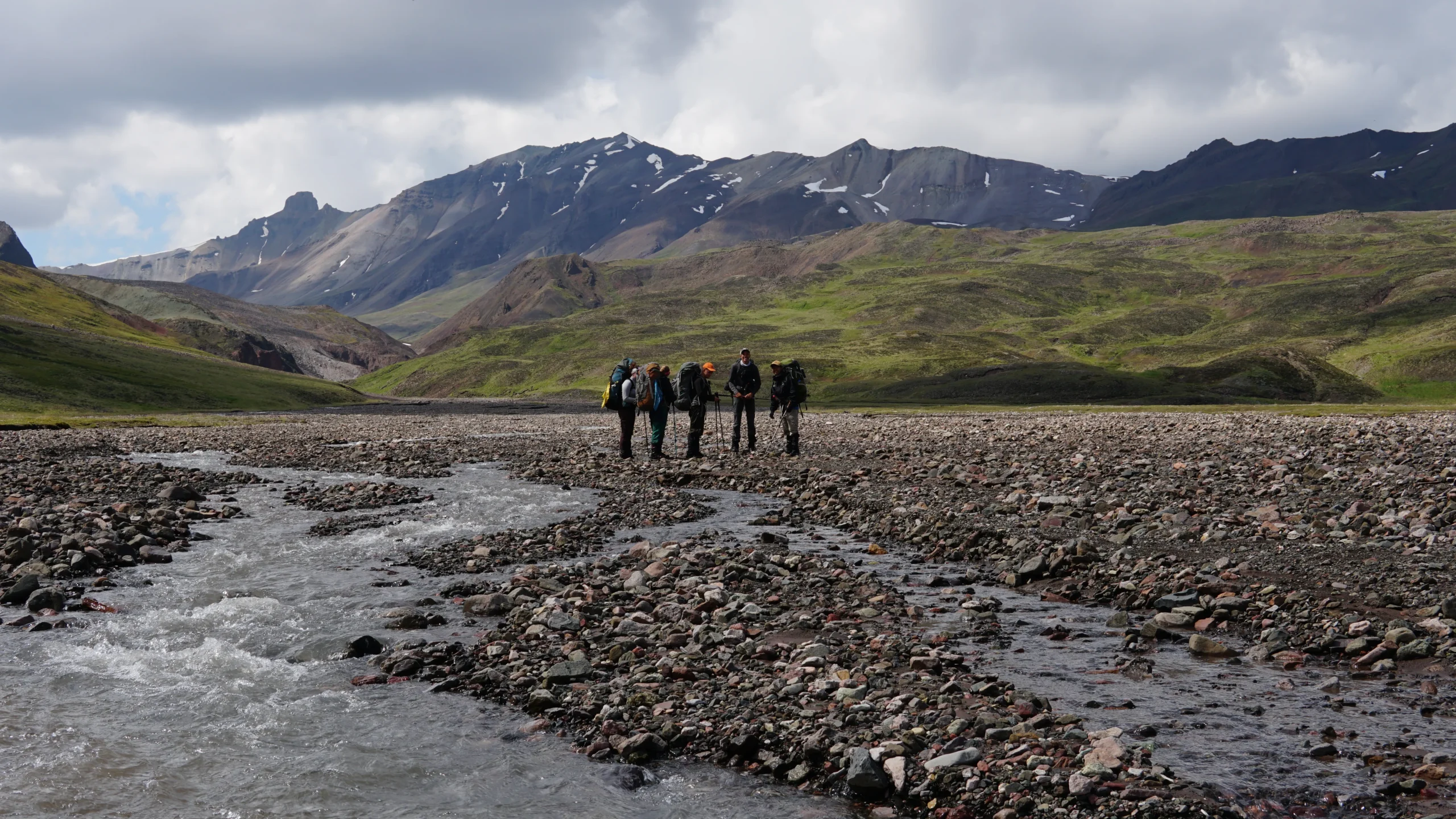 a group of five people stand in a braided stream channel. there are mountains and green hillsides in the background.