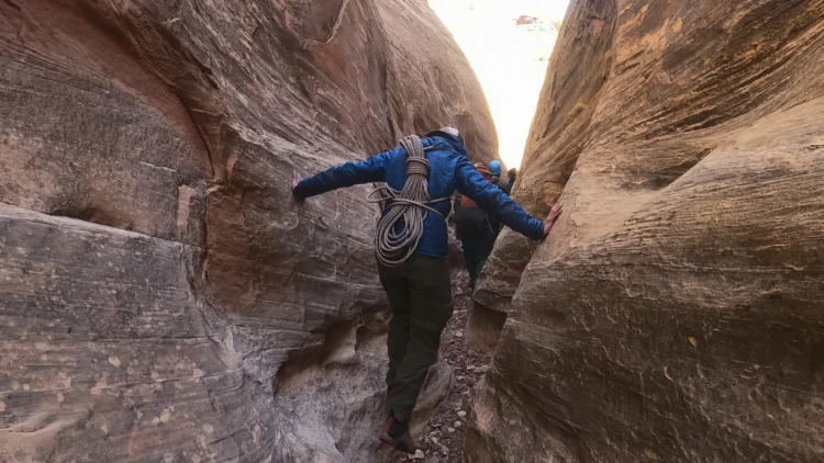a person with a blue jacket carries a climbing rope on their back as tey walk between two narrow and steep slot canyon walls. 