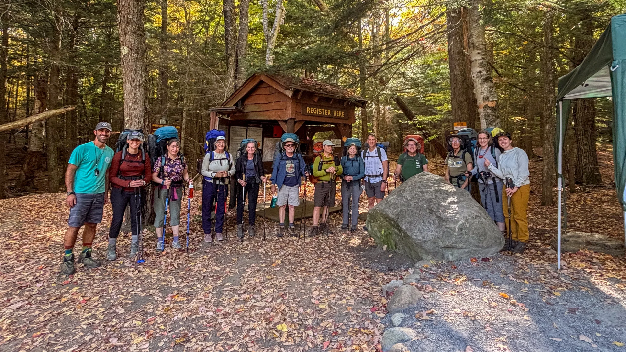 A group of adults pose in a forest by a trail head sign. They are all wearing backpacks.