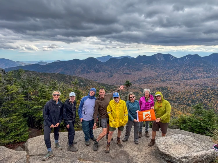 A group of adults in wind and rain gear pose with funny faces on a rocky mountain top. There are more mountains in the background.