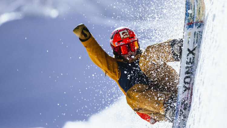 a person in a yellow snowsuit wearing a red helmet with a NOLS logo and riding a snowboard gets big air