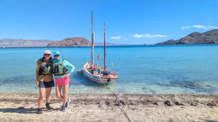 Sailors pose on the beach with the boat anchored in the water behind them