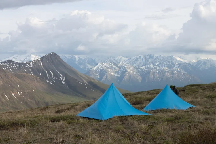 two blue tents site on the tundra. There are low clouds hovering over snow-capped mountains in the distance. 