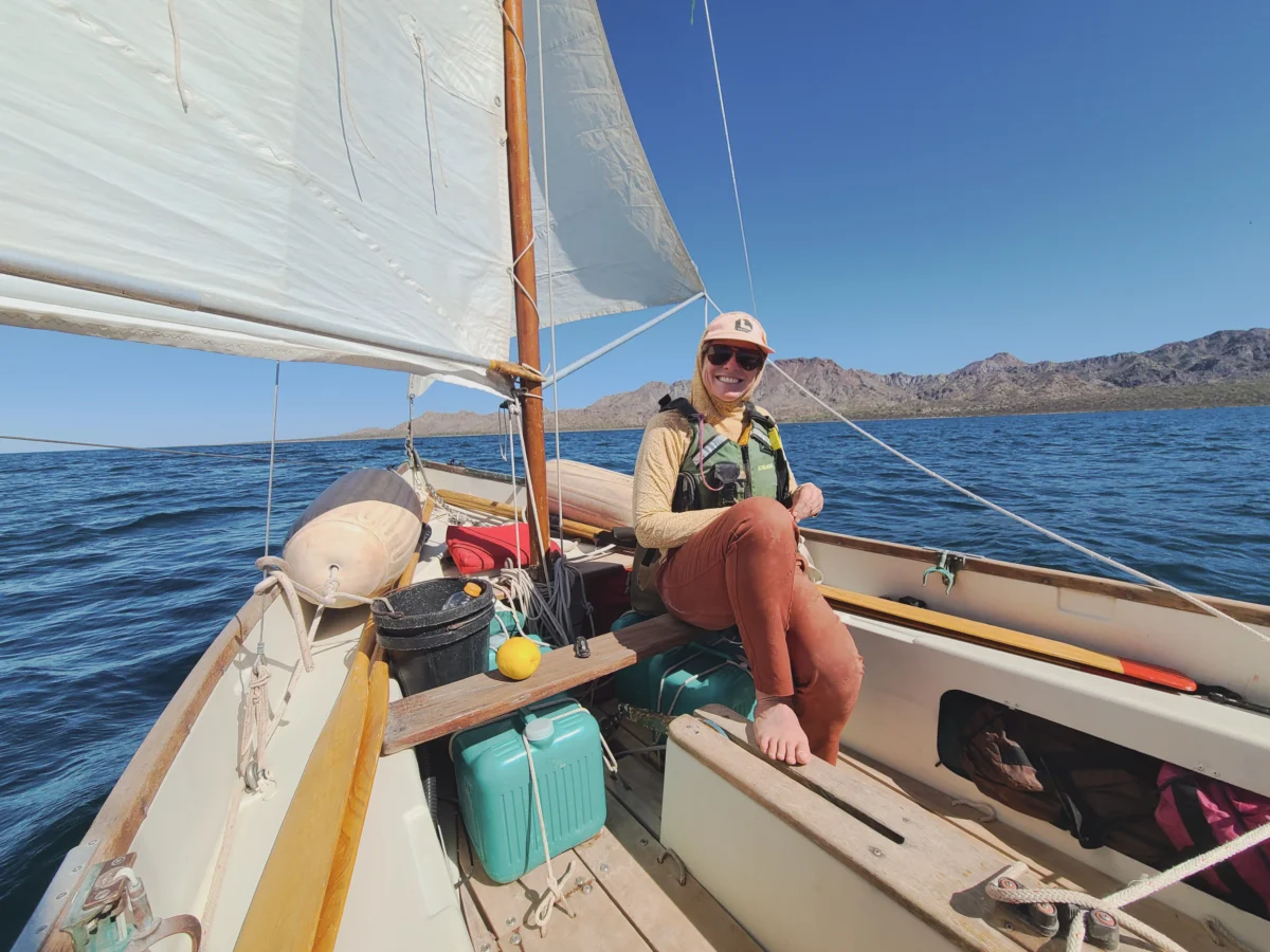 Sailor smiles next to the sail with blue water and skies and mountains in the background