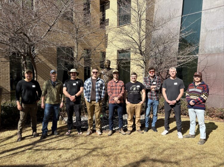 nine smiling men stand together in front of a brick building
