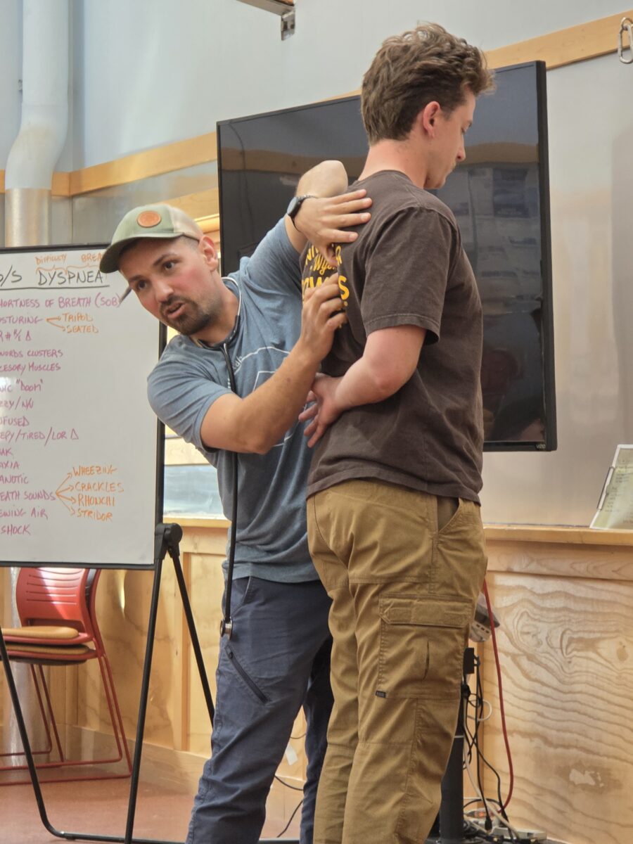 a man in a baseball cap talks to people offscreen while pointing to part of another man's spine.