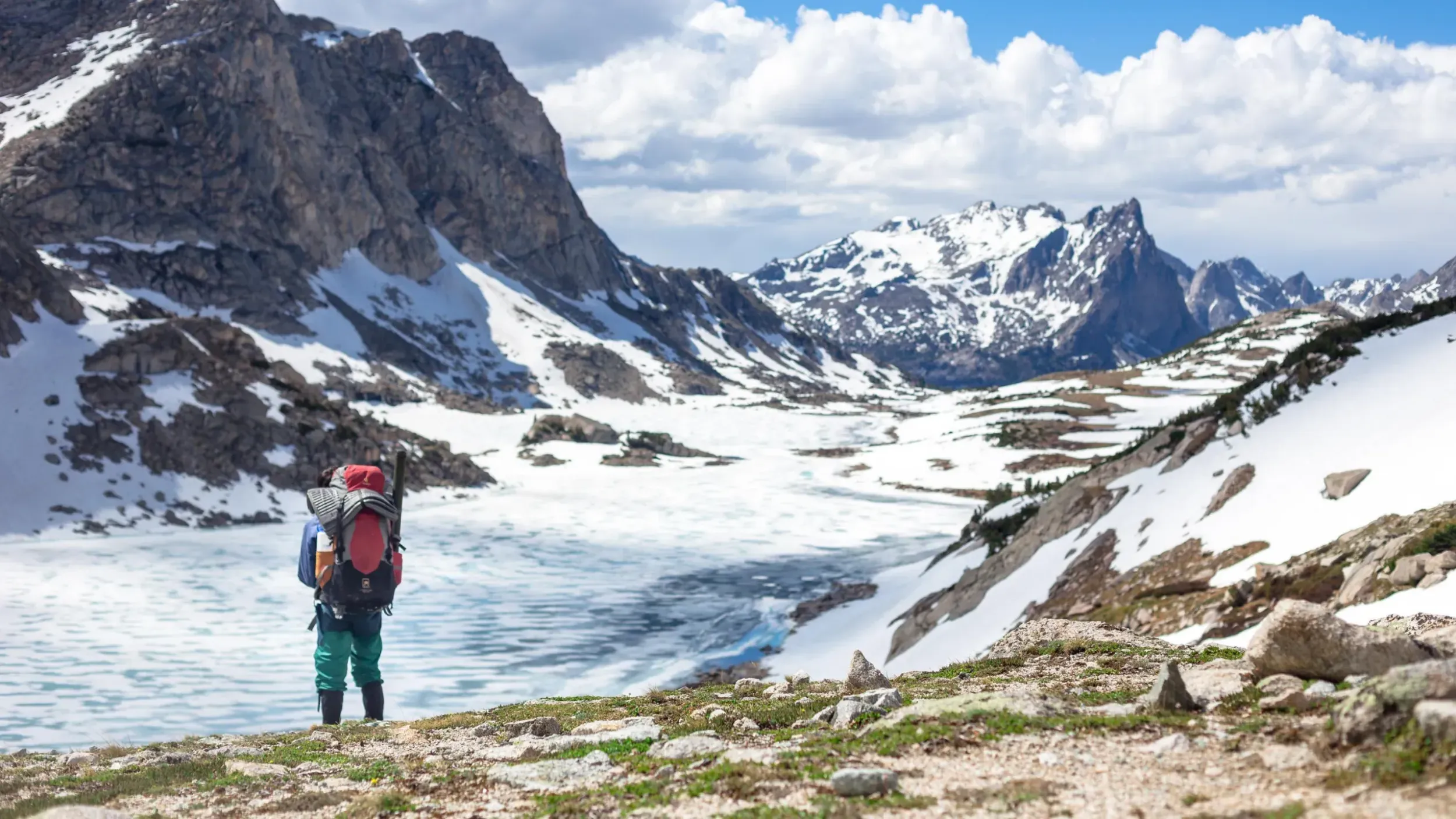 a person wearing a large backpack looks across a frozen lake and steep mountains in the distance