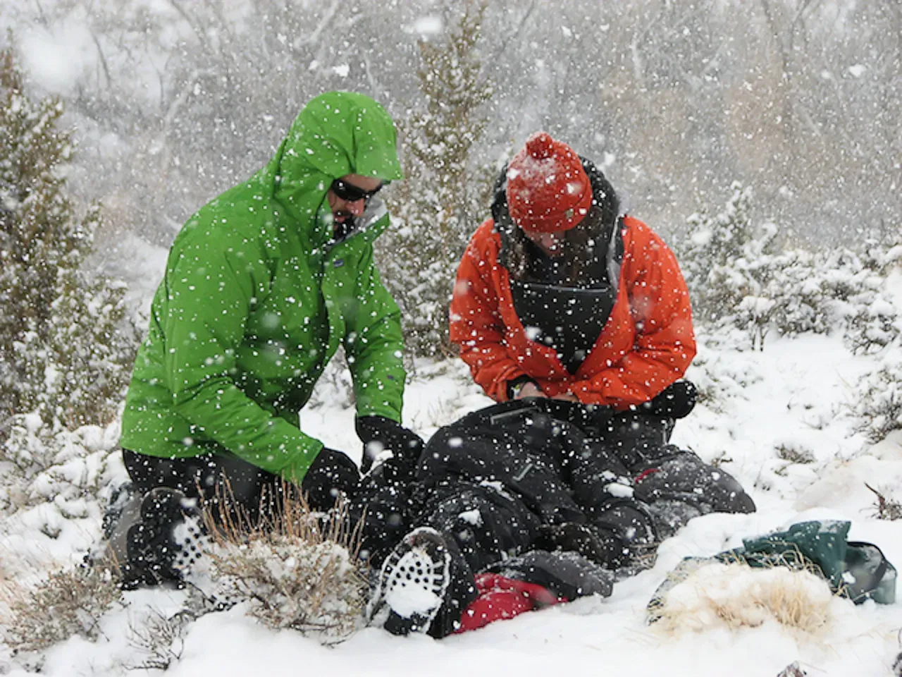two people in winter clothing help a person laying on the ground. it is snowing